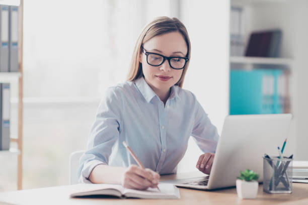 Portrait of charming director, pretty, nice woman holding pencil in hand, writing a date of conversation in reminder, organizing her day, sitting in work place, station at desktop stock photo
