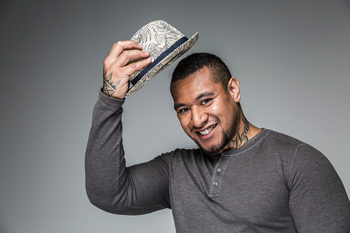 SYD11207: Portrait of smiling young adult pacific islander man with tattoos tipping hat as greeting and respect