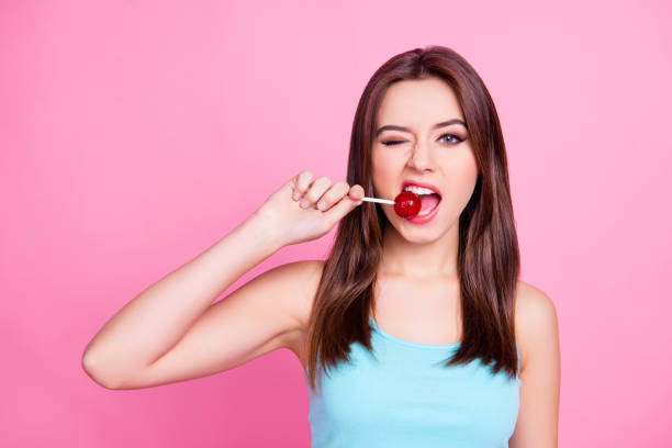 Portrait of lovely sweet cute charming beautiful cheerful hungry adorable gorgeous woman with straight brown hair trying to bite red lollipop  on stick, isolated on bright pink background Portrait of lovely sweet cute charming beautiful cheerful hungry adorable gorgeous woman with straight brown hair trying to bite red lollipop  on stick, isolated on bright pink background candy in mouth stock pictures, royalty-free photos & images