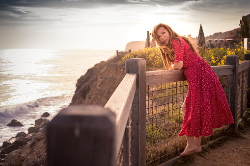 An Asian woman is standing barefoot, enjoying the view of the Pacific Ocean. She's a tourist in an affluent area in Southern California.  A wind is blowing her red dress.  Yellow flowers add to the Spring scene.