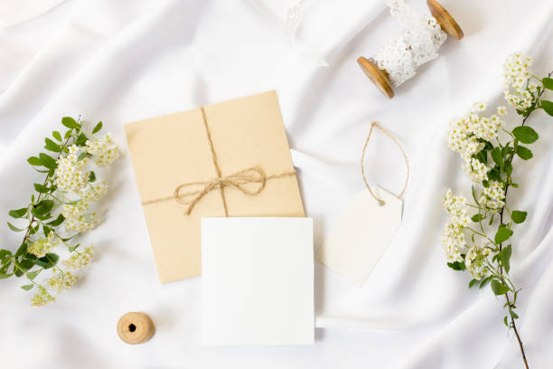 istock Flat lay workspace, mockup. Wedding invitation cards, craft envelopes,white flowers, green leaves and lace on white satin background. Top view 962452804