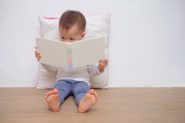 Cute little Asian 18 months / 1 year old toddler boy child sitting on floor, leaning against pillow, looking at a book stock photo