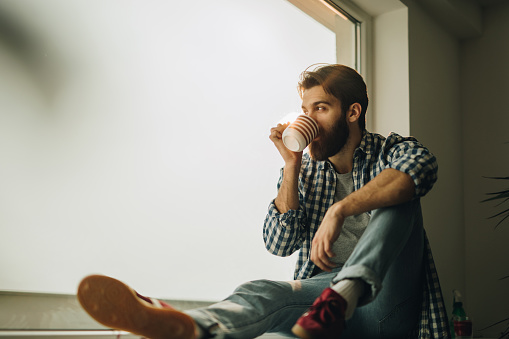 Bearded man drinking coffee by the window and looking through it. Copy space.