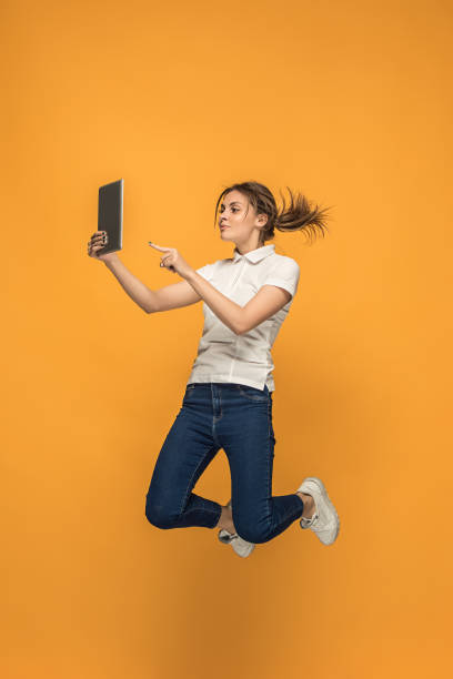 istock Image of young woman over orange background using laptop computer or tablet gadget while jumping 962465910