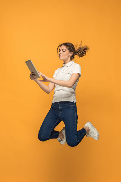 istock Image of young woman over orange background using laptop computer or tablet gadget while jumping 962466004