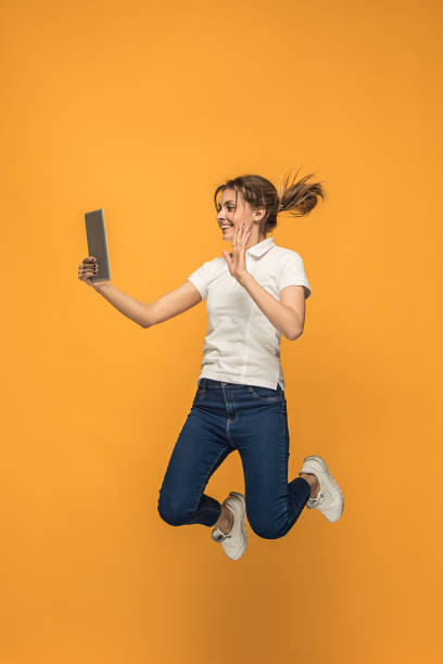 istock Image of young woman over orange background using laptop computer or tablet gadget while jumping 962466058