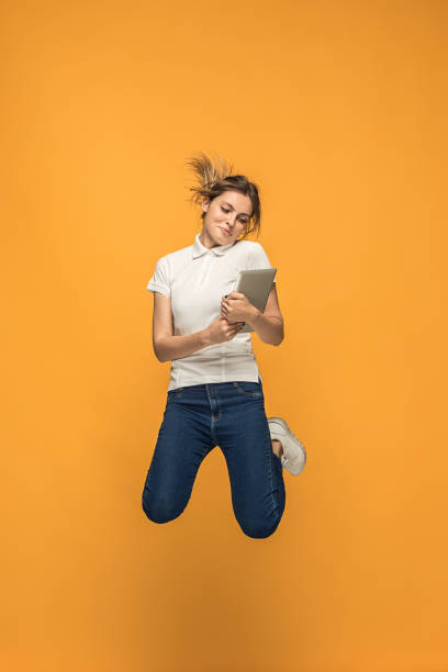 Image of young woman over orange background using laptop computer or tablet gadget while jumping