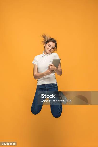 Image Of Young Woman Over Orange Background Using Laptop Computer Or Tablet Gadget While Jumping Stock Photo - Download Image Now