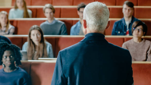 istock Camera Facing Class: College Professor Gives a Lecture to a Classroom Full of Multi Ethnic Students. Talented Speaker Captures Audience Attention. 962475982