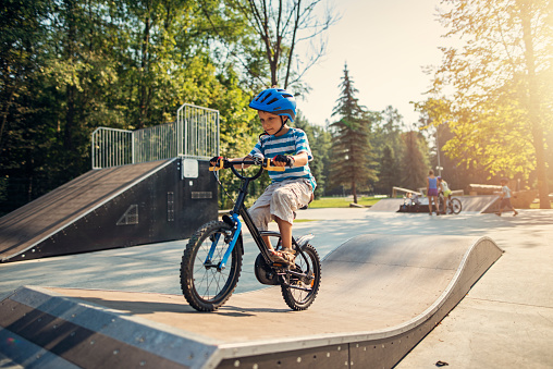 Little boy wearing a helmet is riding a bicycle on ramp in city park on summer day. The boy is aged 6 and is smiling.\nNikon D800