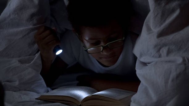 niño leyendo libros en la noche debajo de la manta, él mismo la iluminación con linterna - child bedtime imagination dark fotografías e imágenes de stock