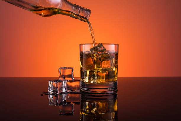 istock Whisky pouring into glass with ice drink on dark orange background 962486614