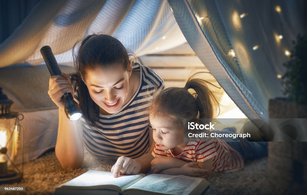 Mom and child reading book Family bedtime. Mom and child daughter are reading a book in tent. Pretty young mother and lovely girl having fun in children room. Flashlight Stock Photo