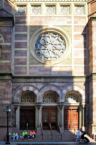 Workers sat on the steps of the Central Synagogue on Lexington Avenue in New York. There are tourist walking past and there is  shadow cast across the image from late afternoon sun.