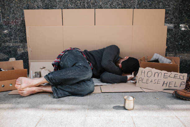 istock Helpless and defenceless man is lying on the cardboard on concrete floor and sleeping. He is covering his face with hands hiding it from the sun. There is a sign besides him says homeless please help 962517616