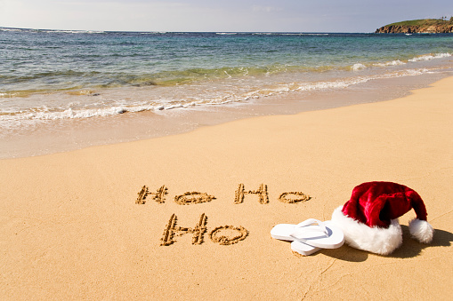 Santa cap and flip flops and a happy Ho Ho Ho written on a sandy beach with waves rolling inward