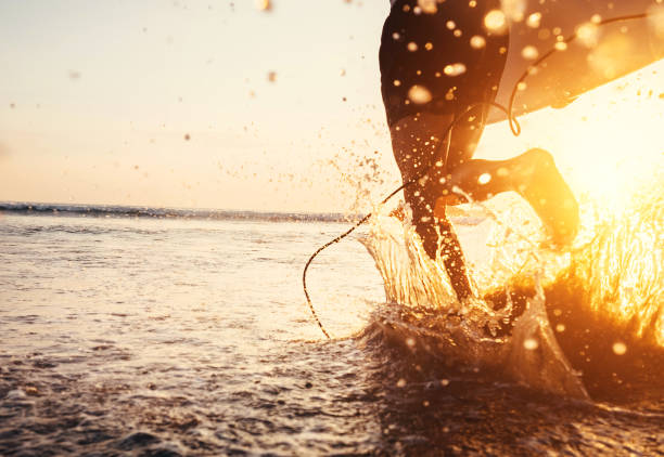 istock Man surfer run in ocean with surfboard. Closeup image water splashes and legs, sunset light 962567960