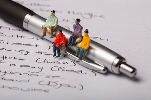 Miniature people figurines sit on a pen which is on a page of hand written words