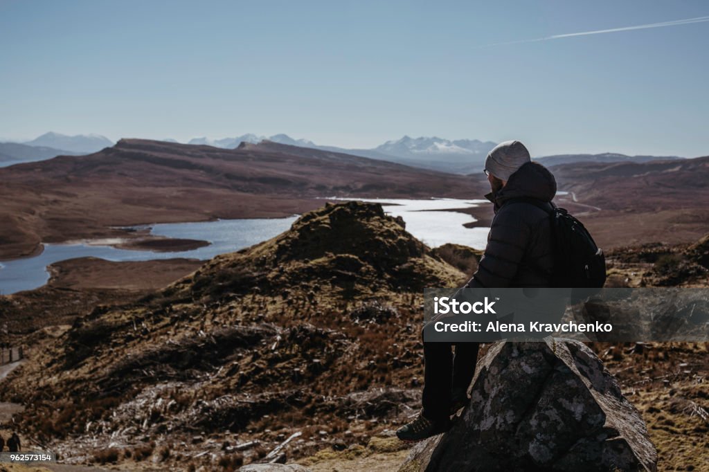 Man relaxing on top of the Old Man of Storr walk on Isle of Skye, Scotalnd, sitting on a stone. Man relaxing on top of the Old Man of Storr walk on Isle of Skye, sitting on a stone, mountains and lake on the background. Adult Stock Photo