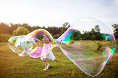 Little girl making a big soap bubble. Focus on the soap bubble. Summer meadow.