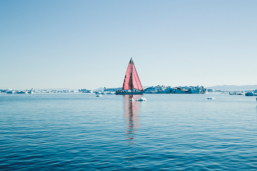 People enjoying beauty of nature by sailboat with red sail on Disko bay near the small town of Ilulissat in Greenland.