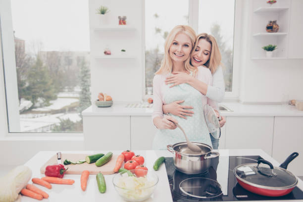 istock Cheerful charming hungry daughter hugging her mother while preparing tasty dinner standing in modern white cook room having vegan products vegetables on the table using kitchenware 962616068