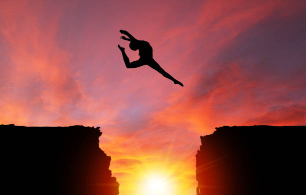 istock Silhouette of Girl Leaping Over Cliffs With Sunset Landscape 962619950