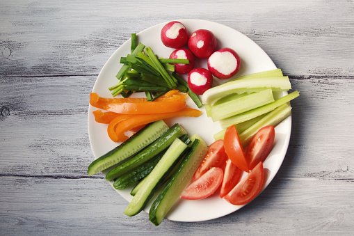 White plate with vegetables for a vegetarian salad. Radishes, tomatoes, celery, bell pepper, onion and cucumber. White wooden kitchen table. Close up. Top view.