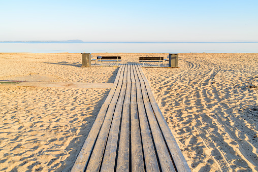 Wooden walkway at a beach, with two empty benches and trash bins on the sides. Windless sea under the horizon on a sunny morning before the bathers arrive. Angelholm in Sweden.