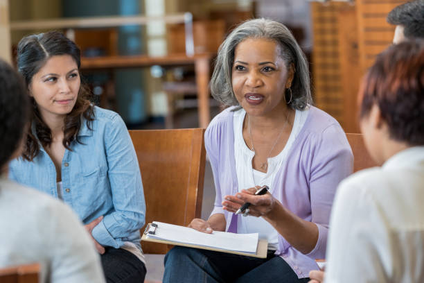 Senior mental health professional facilitates support group Serious senior African American female mental health profession gestures while talking with clients in a support group or group therapy session. She is holding a clipboard. group therapy photos stock pictures, royalty-free photos & images