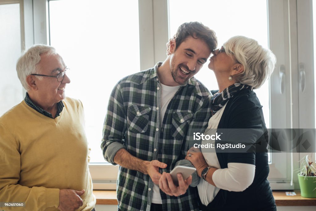 Son with his parents Young man looking at phone, his mother kissing him and father looking at them Parent Stock Photo