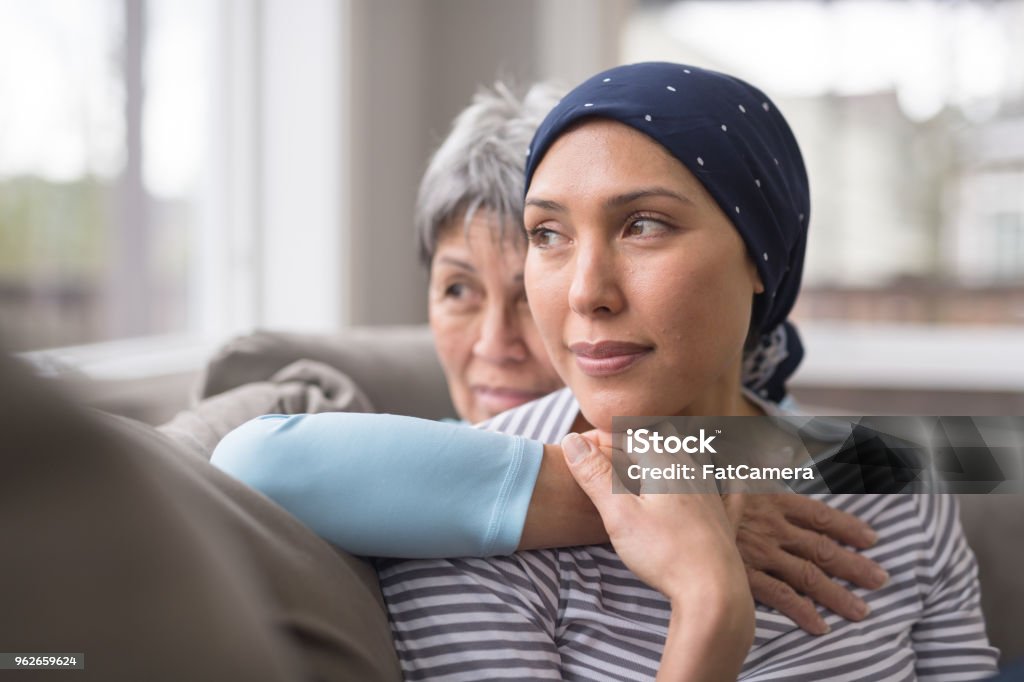 An Asian woman in her 60s embraces her mid-30s daughter who is battling cancer An ethnic woman wearing a headscarf and fighting cancer sits on the couch with her mother. She is in the foreground and her mom is behind her, with her arm wrapped around in an embrace, and they're both looking out the window in a quiet moment of contemplation. Cancer - Illness Stock Photo