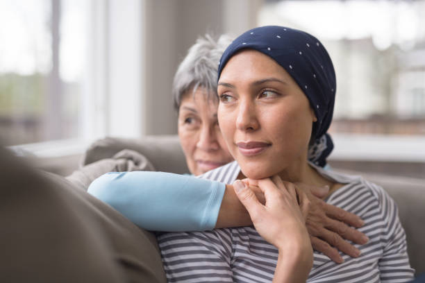 An Asian woman in her 60s embraces her mid-30s daughter who is battling cancer An ethnic woman wearing a headscarf and fighting cancer sits on the couch with her mother. She is in the foreground and her mom is behind her, with her arm wrapped around in an embrace, and they're both looking out the window in a quiet moment of contemplation. determination asian stock pictures, royalty-free photos & images