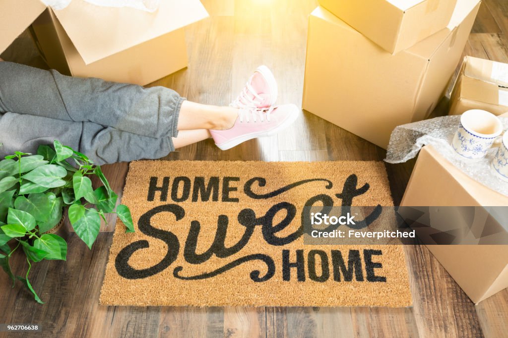 Woman Wearing Sweats Relaxing Near Home Sweet Home Welcome Mat, Moving Boxes and Plant. Moving House Stock Photo