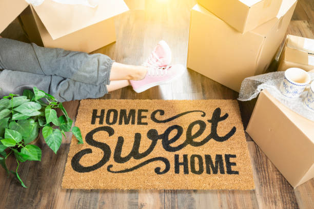 Woman Wearing Sweats Relaxing Near Home Sweet Home Welcome Mat, Moving Boxes and Plant. stock photo