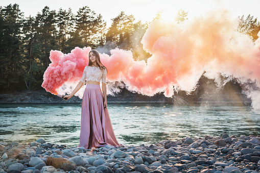 girl with a smoke bomb on the bank of a mountain river with pink and red smoke and forest against a background