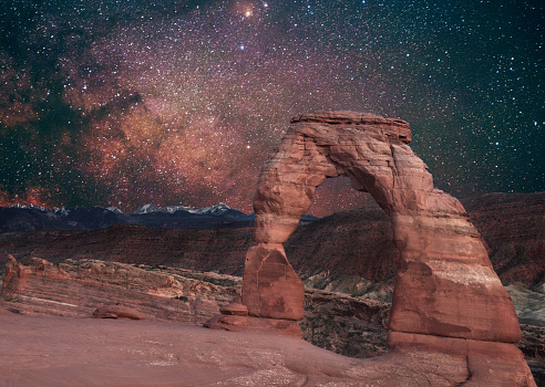 This is a color photograph of landmark, Delicate Arch in Arches National Park in Utah, USA at night with the Milky Way in the sky. The high desert landscape is full of rock formations created by erosion in sandstone over time.