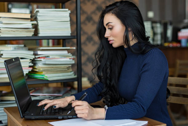 istock Side view. Young business woman sitting at table and taking notes in notebook. On table is laptop. Student learning online. Blogger 962754770