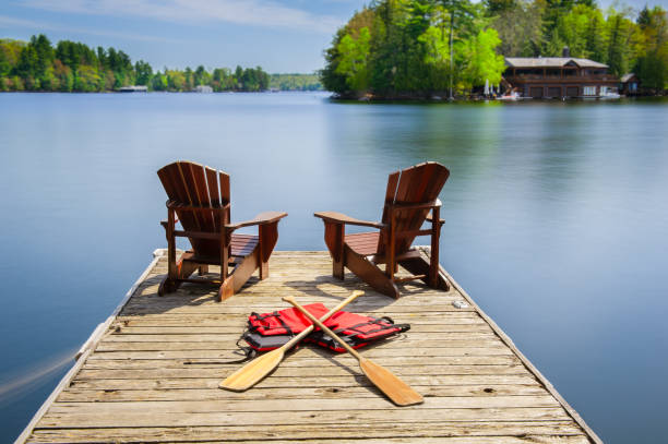 Muskoka chairs on a wooden dock Two Muskoka chairs on a wooden dock facing a lake. Paddles and life jackets are visible on the dock. Across the calm water there is a brown cottage. calm water photos stock pictures, royalty-free photos & images