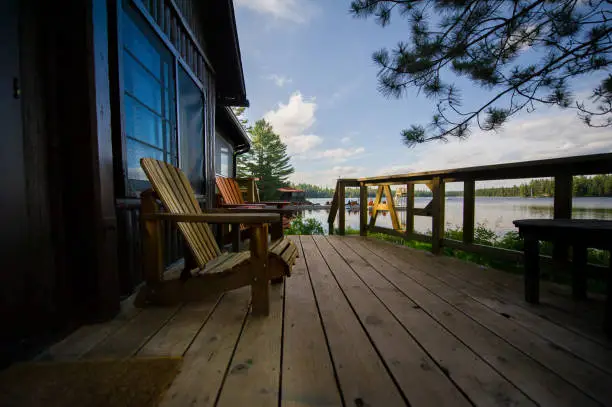 Two Muskoka chairs on a wooden deck facing a lake. In the background there’s a pier with a big amount of chairs