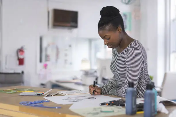 A graphic designer sketches out prototypes for an idea onto paper. She is using a pen and leaning over her desk to concentrate. There are various pens, papers, and art supplies setting on the desk. Light is coming in from large exterior windows in the background.