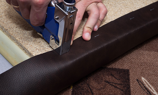 Craftsman fastening brown leather to the paricle board using staples and blue stapler