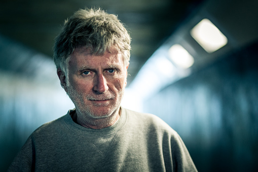 Close up view of a homeless senior adult male, of caucasian ethnicity, standing outdoors in a dark, cold subway tunnel. The man, who has white hair and white stubble on his face, has a hopeless and sad expression on his face. Behind the man, the dark tunnel recedes into the distance, out of focus. Lots of room for copy space.