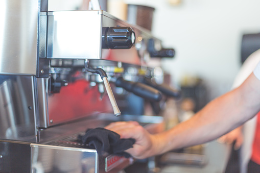 A barista wipes down a stainless steel espresso machine that is on the counter of a coffee shop. The shot is focused tight on the machine.
