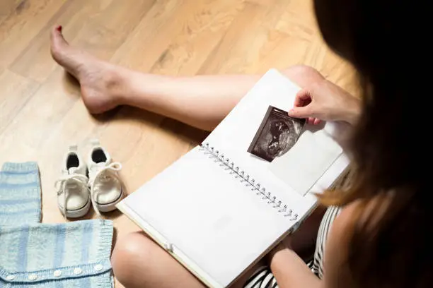 Woman placing baby's sonogram into baby's first year memory book. Baby clothes and sneakers laying on the floor