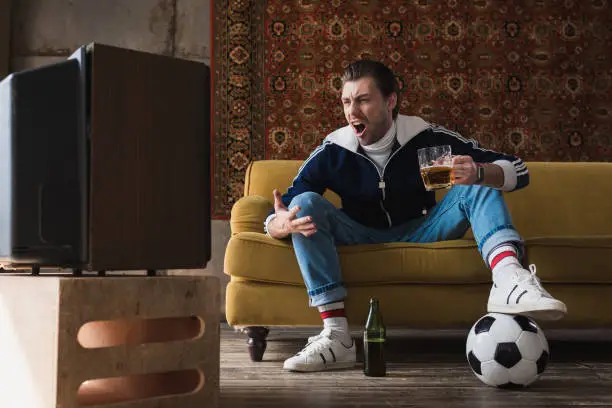 expressive young man in vintage clothes with ball and mug of beer watching soccer on old tv and shouting