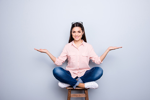 Portrait of pretty charming trendy woman sitting on wooden bar chair with crossed legs holding two copy spaces empty places on her palms looking at camera isolated on grey background