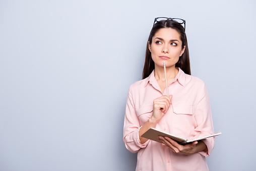 Portrait with copyspace of thoughtful concentrated busy charming woman having notepad and pen in hands planning, expertising, analyzing isolated on grey background, advertisement concept