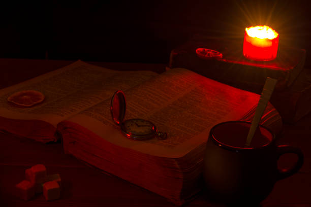 istock Old book on the table. The open Holy Bible is on the table. Ancient key. A burning candle over the Bible illuminates its pages. Mug with a spoon, pieces of sugar and orange slices. 962925886