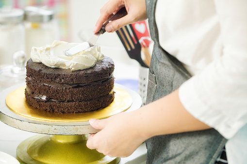 Close-up on baker's hands spreading butter cream icing on layered Chocolate cake using a spatula